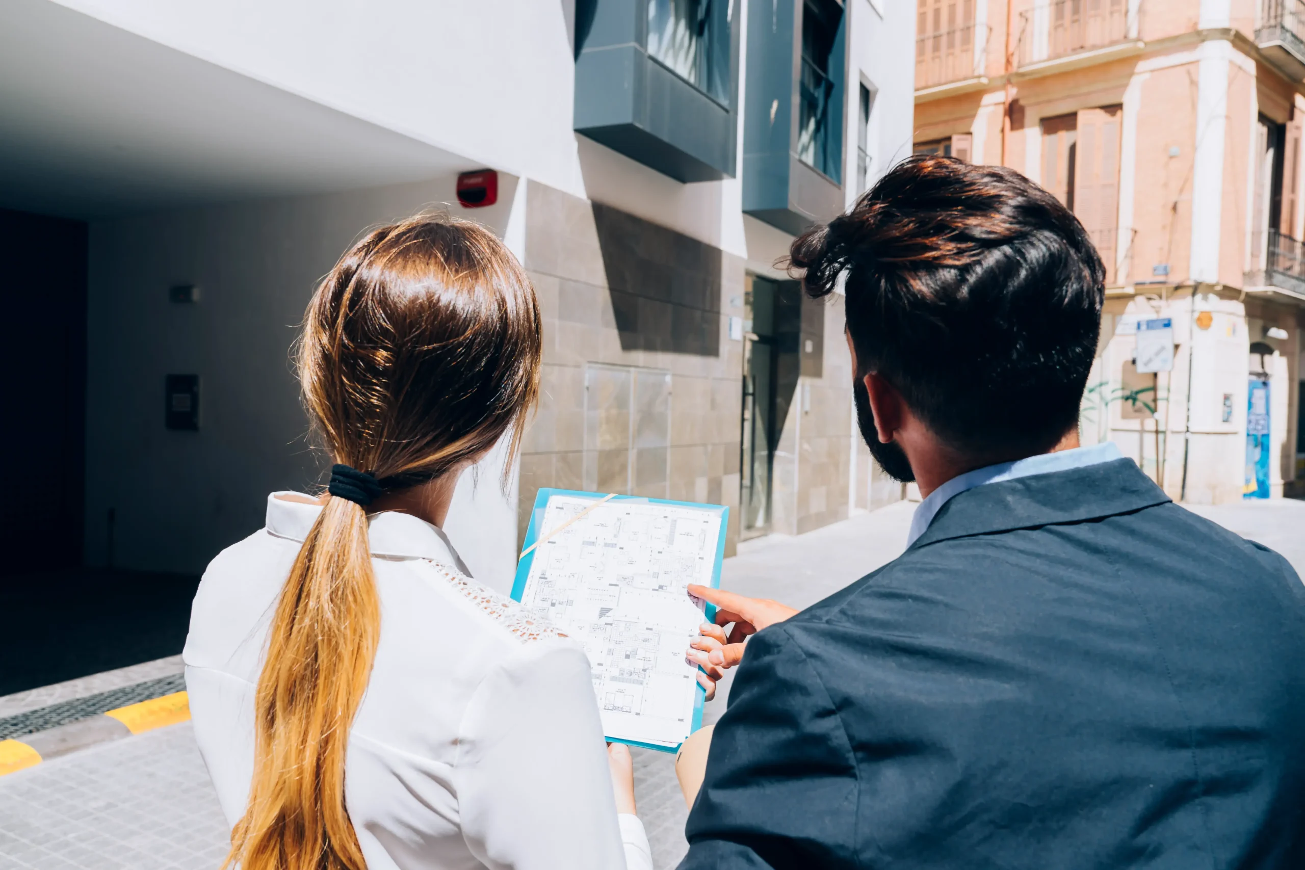 Two professionals stand outside a modern building. One holds a blueprint or map while the other points at it, suggesting they’re reviewing property details, site plans, or evaluating a location for real estate or construction.